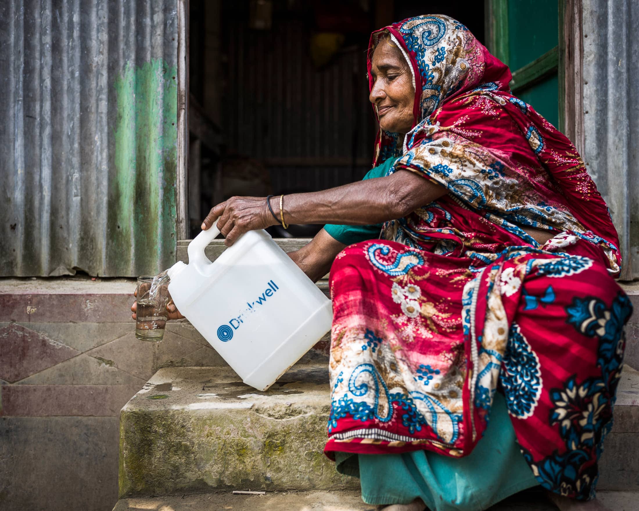 A woman from Bangladesh pouring a glass of water with a canister of water branded Drinkwell 一位来自孟加拉国的妇女用一罐标有Drinkwell的水倒了一杯水